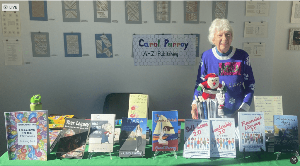 Author in hiliday sweater before pale wall with small artwork on it, plus sign: Carol Purroy, A-Z Publishing, on it. Table full of books ar forefront.